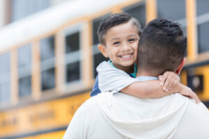 A confident little boy smiles for the camera as he embraces his father after the first day of school. He has just ridden the school bus home.